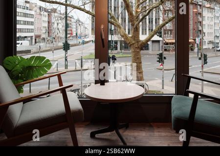 Ein stilvoller Café-Sitzbereich mit einem runden Tisch und zwei modernen Sesseln, die durch ein großes Fenster mit Blick auf eine geschäftige Stadtstraße in Düsseldorf angeordnet sind. Stockfoto