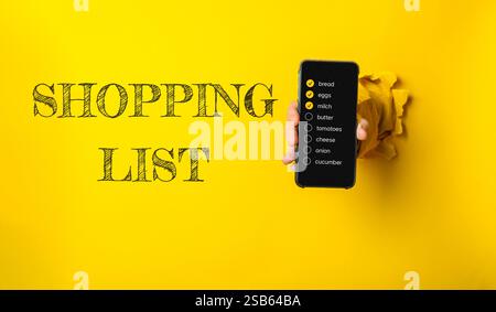 Person, die ein Handy mit einer Einkaufsliste hält. Die Einkaufsliste ist auf gelbem Hintergrund Stockfoto
