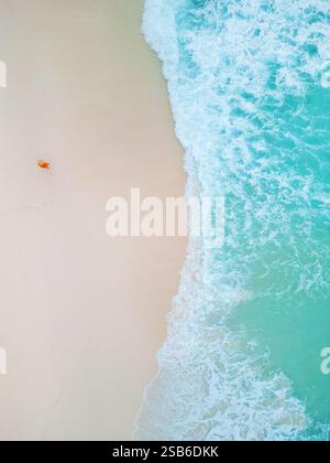 Luftaufnahme einer Frau, die an einem weißen Sandstrand entlang spaziert und Spuren in der Nähe der türkisfarbenen Meereswellen in einer ruhigen und isolierten Küstenszene hinterlässt Stockfoto
