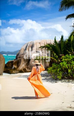 Eine Frau in orangefarbenem Kleid und Sonnenhut spaziert an einem Sandstrand auf den Seychellen, umgeben von Palmen und Granitfelsen unter einem hellblauen Himmel Stockfoto
