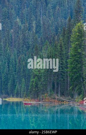 Ein einsamer Fischer in einem roten Boot, der friedlich auf dem ruhigen Smaragdsee fischt, umgeben von üppigen immergrünen Wäldern. Stockfoto