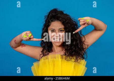 Lächelnde junge Frau mit lockigen Haaren und gelben Nägeln, die mit beiden Händen auf blauem Hintergrund nach unten zeigen Stockfoto