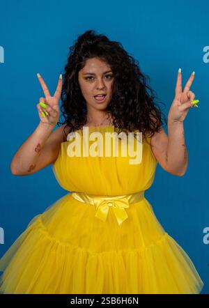 Stilvolle junge Frau, die mit beiden Händen Frieden spendet, in gelbem Kleid auf blauem Hintergrund Stockfoto
