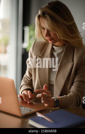 Die junge, schöne blonde Frau arbeitet in einem hellen und stilvollen Büro. Sie tippt auf ihrem Laptop, während sie Notizen in einem Notizbuch macht. Stockfoto