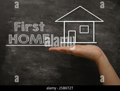 Hand hält eine Tafel mit den Worten First Home drauf. Das Schwarze Brett ist mit weißer Kreide bedeckt, und die Hand hält es hoch, um den zu zeigen Stockfoto