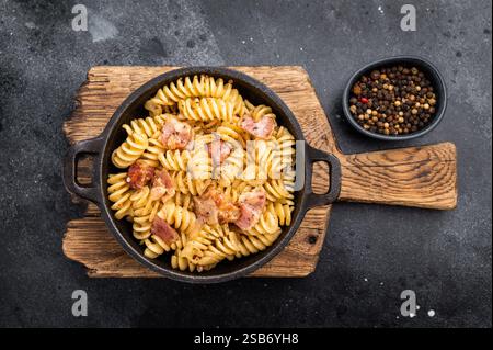 Fusilli Carbonara Nudeln mit knusprigen Speckstückchen und Parmesankäse in einer Pfanne. Schwarzer Hintergrund. Draufsicht. Stockfoto