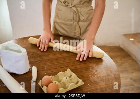Person mit beigefarbener Schürze, die Teig auf einem Holztisch rollte. Zutaten wie Eier und Mehl sind in der Nähe zu sehen und schaffen eine warme und gemütliche Atmosphäre Stockfoto
