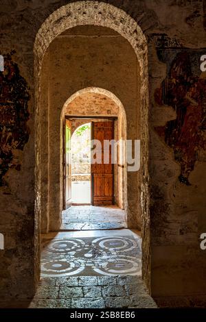 Ein historischer Steinbogen im Kloster Andromonastiro in Messinia, Griechenland, mit einer offenen Holztür, die zu einem sonnendurchfluteten Innenhof führt. Stockfoto