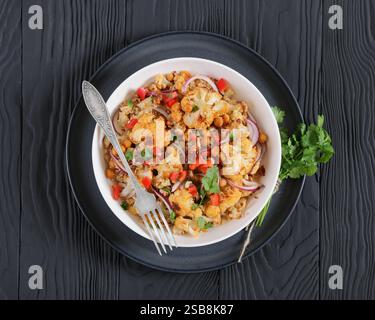 Gebratener Blumenkohl-Quinoa-Salat mit Walnüssen, rotem Pfeffer, Zwiebeln und Koriander in weißer Schüssel mit Gabel auf schwarzem Holztisch, horizontale Ansicht, Stockfoto