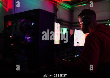 Junger Mann mit Brille und Kopfhörern, der in einem Computerspielzimmer sitzt und sich auf einen Bildschirm konzentriert, mit farbenfrohen Neonlichtern im Hintergrund. Stockfoto