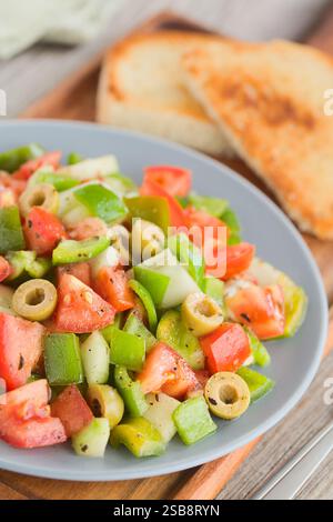 Frischer veganer Salat aus Tomaten, Oliven, grünem Pfeffer und Gurke, gewürzt mit Oregano, Salz, schwarzem Pfeffer und Olivenöl, serviert auf blauem Teller Stockfoto