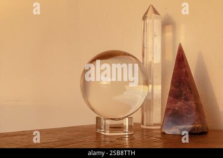Klare Kristallkugel, Quarzspitze und Amethyst-Pyramide auf einer Holzoberfläche, in warmes Sonnenlicht getaucht. Stockfoto