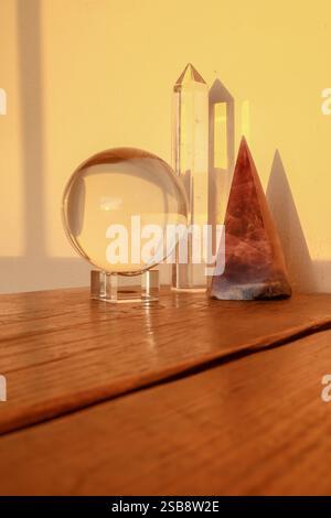 Klare Kristallkugel, Quarzspitze und Amethyst-Pyramide auf einer Holzoberfläche, in warmes Sonnenlicht getaucht. Stockfoto