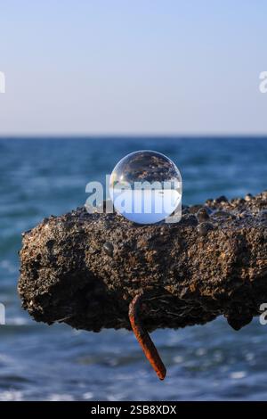 Der Lensball sitzt auf einem rauen, felsigen Felsvorsprung am Meer und spiegelt Himmel und Meer wider Stockfoto