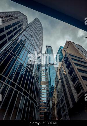 Flacher Blick auf die hoch aufragenden Wolkenkratzer in Hongkong, deren Glasfassaden den bewölkten Himmel reflektieren. Der städtische Canyon schafft einen dramatischen, aufwärts gerichteten pro Stockfoto