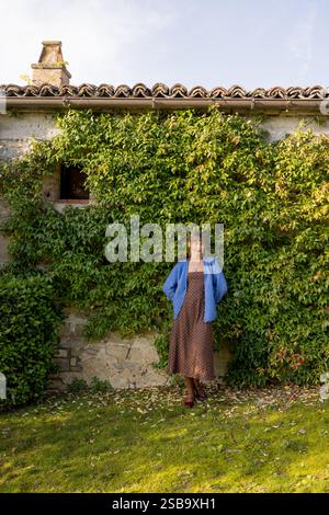 Eine stilvolle Frau in einem blauen Strickjacke und einem Kleid mit Punktmuster steht selbstbewusst neben einer rustikalen Steinwand, die mit leuchtendem grünem Efeu geschmückt ist. Die ruhige Umgebung im Freien spiegelt den friedlichen ländlichen Charme wider Stockfoto
