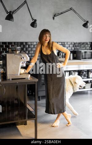Eine barfüßige Frau in einem minimalistischen Kleid lehnt sich auf eine Kaffeemaschine und lächelt in einer stilvollen Küche mit dunklen Fliesen. Ihr weißer Hund geht vorbei und sorgt für Wärme in der gemütlichen und eleganten Umgebung Stockfoto