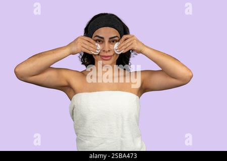 Die junge Frau, die in ein Handtuch gewickelt ist, reinigt ihr Gesicht mit Wattepads auf violettem Hintergrund Stockfoto