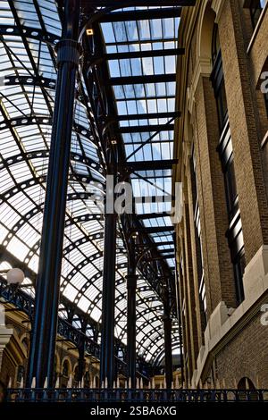 Eine perspektivische Aussicht auf eine Dachkonstruktion mit Glas- und Metallrahmen über einem Backsteingebäude. Dunkle Metallsäulen und -Träger tragen das gewölbte Glas ce Stockfoto