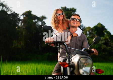 Ein lächelndes Paar fährt mit dem Motorrad durch üppig grüne Reisfelder. Die Frau umarmt den Mann von hinten, während sie auf dem Motorrad fahren. Erkunden Sie die malerische Landschaft Stockfoto