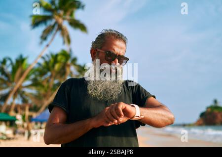 Ein älterer, stilvoller Mann mit grauem Bart benutzt am Strand eine intelligente Uhr. Trendiger reifer Mann in Sonnenbrille kariert tragbares Gadget. Aktiver Lebensstil, technisch versiert Stockfoto