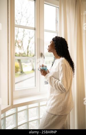 Eine junge Frau in bequemer Loungewear, die einen Becher hält, steht an einem hellen Fenster und blickt nachdenklich auf die Landschaft im Freien, während sie ihren Morgen genießt Stockfoto