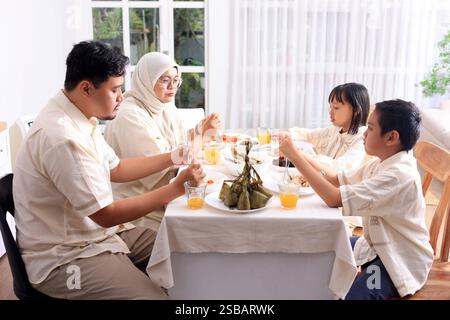 Junge asiatische muslimische Familie, Vater, Mutter und zwei Kinder essen Ketupat Lebaran zusammen, feiern Idul Fitri Eid Al Fitr Stockfoto