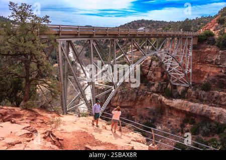 Ein Paar fotografiert eine Brücke über einen Fluss. Die Brücke ist sehr hoch und überspannt eine lange Strecke. Das Paar steht auf einem felsigen Hügel. Stockfoto