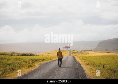 Alleinreisende spazieren auf einer ruhigen Straße in Richtung Ingjaldsholskirkja Kirche auf der Halbinsel Snaefellsnes, Island. Umgeben von goldenen Feldern unter einem so Stockfoto