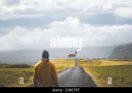 Alleinreisende spazieren auf einer ruhigen Straße in Richtung Ingjaldsholskirkja Kirche auf der Halbinsel Snaefellsnes, Island. Umgeben von goldenen Feldern unter einem so Stockfoto