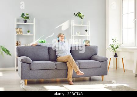 Ältere Frau sitzt zu Hause auf dem Sofa und hört Musik oder Hörbuch mit Kopfhörern Stockfoto