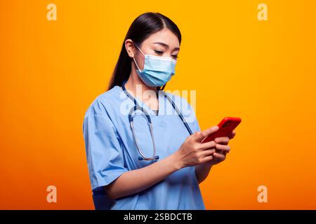Junge Krankenschwester in medizinischer Uniform und Gesichtsmaske, SMS auf ihrem mobilen Gerät, während sie vor isoliertem Hintergrund steht. Asiatische Ärztin mit einem Stethoskop, mit Handy, in ihren sozialen Medien. Stockfoto