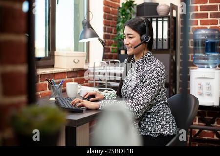 Lächelnde Geschäftsfrau, die auf einer Computertastatur schreibt und mit Kopfhörern lebhafte Gespräche führt. Weibliche Führungskraft setzte sich an ihrem Schreibtisch, benutzte ein Headset und sprach mit Kollegen über einen Anruf. Stockfoto
