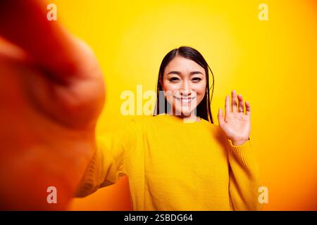 Lächelnde asiatische Dame, die eine Hallo-Geste in Richtung Kamera macht, vloggt für ihr Online-Publikum. Schöne Frau, die mit der Hand winkt und ein Selfie vor gelbem Hintergrund macht, pov-Konzept. Stockfoto