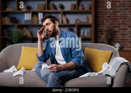 Kranker, kranker Mann, der sich unwohl fühlt, Kopfschmerzen hat, Mann, der auf einer Couch in einem gemütlichen Wohnzimmer sitzt und sich unwohl fühlt, während er Taschentücher hält und seine reibt Stockfoto