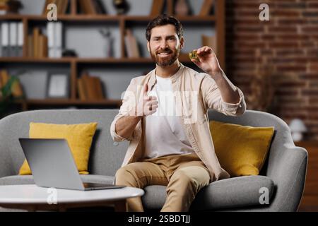 Der Mann lächelt, während er einen Keks hält und in einem gemütlichen Wohnzimmer mit offenem Laptop und heller Einrichtung die Daumen nach oben drückt, was auf einen freudigen Moment hinweist Stockfoto