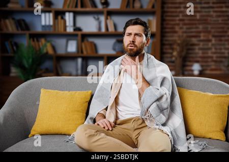 Kranker, kranker Mann, der sich unwohl fühlt, Kopfschmerzen hat, Mann, der auf einer Couch sitzt, die in eine Decke gewickelt ist, mit einem besinnlichen Ausdruck in einem gemütlichen Wohnzimmer Stockfoto