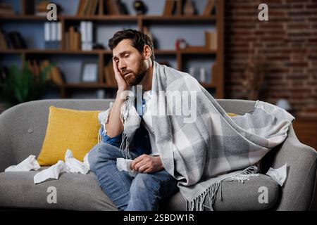 Kranker, kranker Mann, der sich unwohl fühlt, Kopfschmerzen hat, Mann, der auf einer Couch sitzt, die in eine Decke gewickelt ist und Anzeichen von Unwohlsein zeigt, während er von Taschentüchern umgeben ist Stockfoto