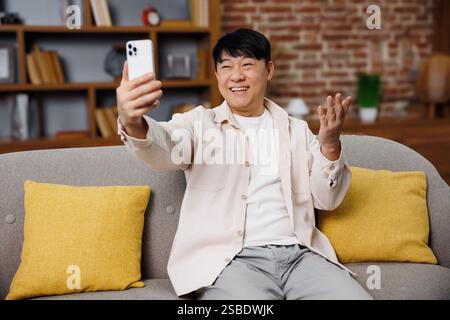 Videogespräche, Telefongespräche, lächelnder asiatischer Mann, genießt Videoanrufe, während er auf einer bequemen Couch in einem gemütlichen Wohnzimmer sitzt Stockfoto