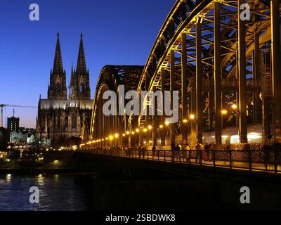 Kölner Dom und Hohenzollernbrücke, abends beleuchtet mit Fußgängern und vorbeifahrendem Zug. Stockfoto