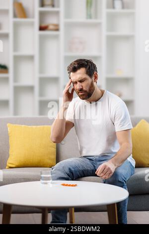 Mann, der Pille nimmt, kranker Mann, hat Kopfschmerzen, Mann sitzt mit schmerzhaftem Ausdruck auf der Couch, hält seinen Kopf, während er auf einem Tisch in Orangentabletten schaut Stockfoto