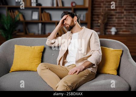 Kopfschmerzen, depressiver Mann, schlechte Gedanken, überwältigt, sitzt ein Mann auf einer grauen Couch in einem gemütlichen Innenraum und legt seinen Kopf in einer Hand Stockfoto