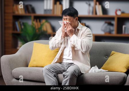 Kranker, kranker Mann, sich unwohl fühlt, Kopfschmerzen hat, asiatischer Mann, der Unbehagen ausdrückt, während er auf einem grauen Sofa in einem gemütlichen Wohnzimmer mit Pflanzen sitzt und Stockfoto