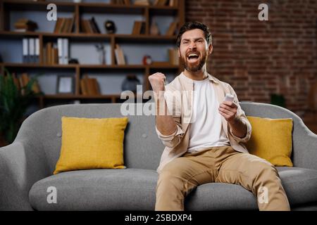 Fans schauen sich ein Spiel an, feiern den Sieg, begeisterte Freunde beim Fernsehen, aufgeregter Mann, der einen Sieg feiert, während er auf einer Couch in einem gemütlichen Wohnzimmer sitzt Stockfoto
