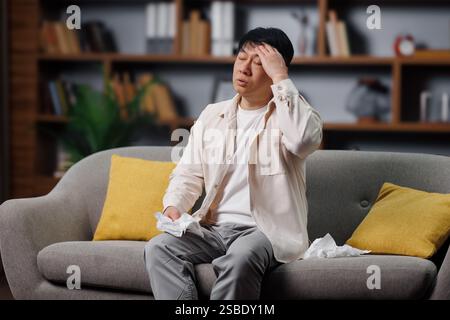 Kranker, kranker Mann, Unwohlsein, Kopfschmerzen haben, Einzelperson, die auf einer Couch sitzt, scheint beunruhigt zu sein, während sie Gewebe hält, was auf einen emotionalen Moment hindeutet Stockfoto