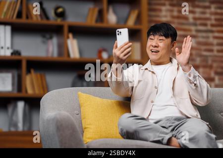 Videogespräche, Telefongespräche, lächelnder asiatischer Mann, auf grauem Sofa glücklich mit Smartphone für Videogespräche in gemütlichem Wohnzimmer Stockfoto
