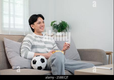 Ein entspannter junger asiatischer Mann isst gerne Popcorn, während er das Spiel seiner Lieblingsmannschaft zu Hause im Fernsehen ansieht. Lifestyle, Freizeit, Sportmeister Stockfoto