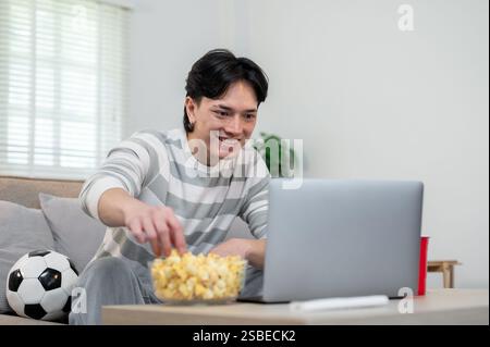 Ein entspannter junger asiatischer Mann isst gerne Popcorn, während er das Spiel seiner Lieblingsmannschaft auf seinem Laptop zu Hause ansieht. Lifestyle, Freizeit, Sport, Ka Stockfoto
