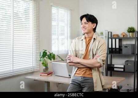 Ein positiver, lächelnder asiatischer Geschäftsmann in lässiger Kleidung steht mit gekreuzten Armen, lehnt sich auf einen Schreibtisch in seinem Büro, träumt oder reflektiert ihn Stockfoto