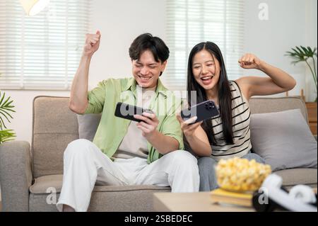 Ein überfröhliches, glückliches asiatisches Paar spielt gerne gemeinsam ein Rennspiel auf ihrem Smartphone und hat eine lustige Zeit zu Hause. Spaß, Wettbewerb, Unterhaltung Stockfoto